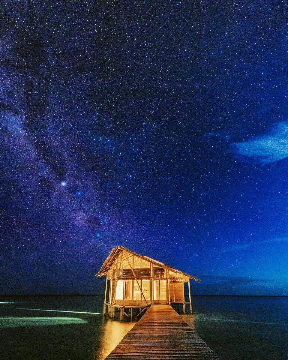 Hình ảnh đẹp 3D về ngôi nhà gỗ phát sáng trên mặt nước dưới bầu trời đầy sao và dải Ngân Hà